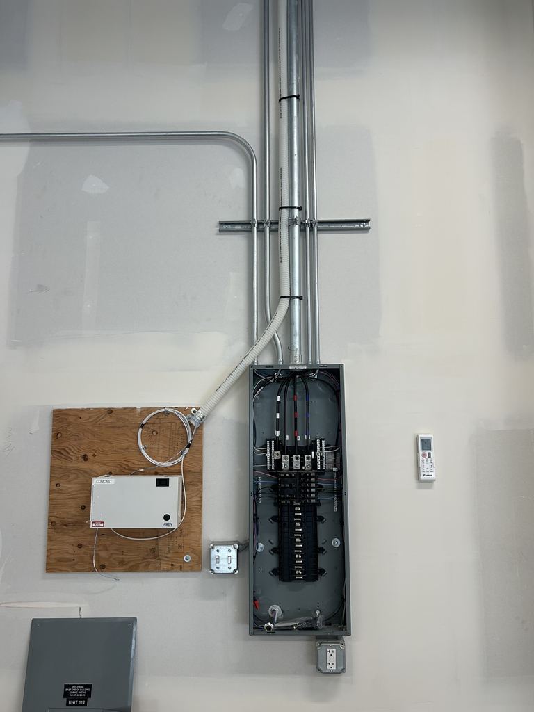 Commercial electrical panel with its cover off, fed by metal conduit running up the wall, beside a telecom box on a plywood backboard