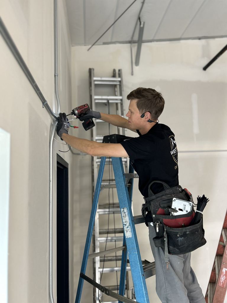 Electrician in a black WestPeak Electric shirt on a blue stepladder, fastening metal conduit to a wall with a drill