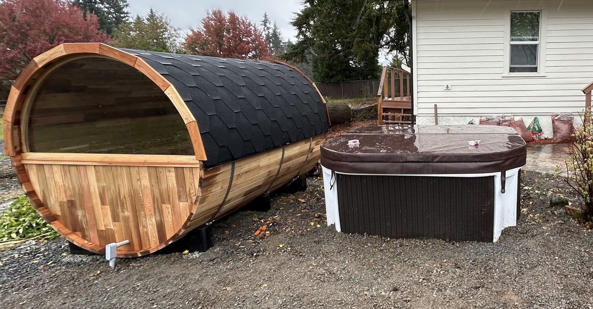 Cedar barrel sauna and a covered hot tub set on gravel beside a house