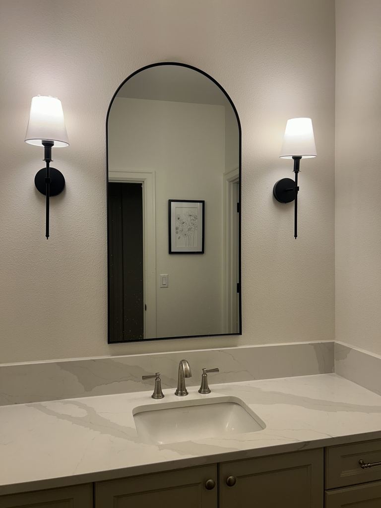 Bathroom vanity with an arched mirror between two black wall sconces with white shades